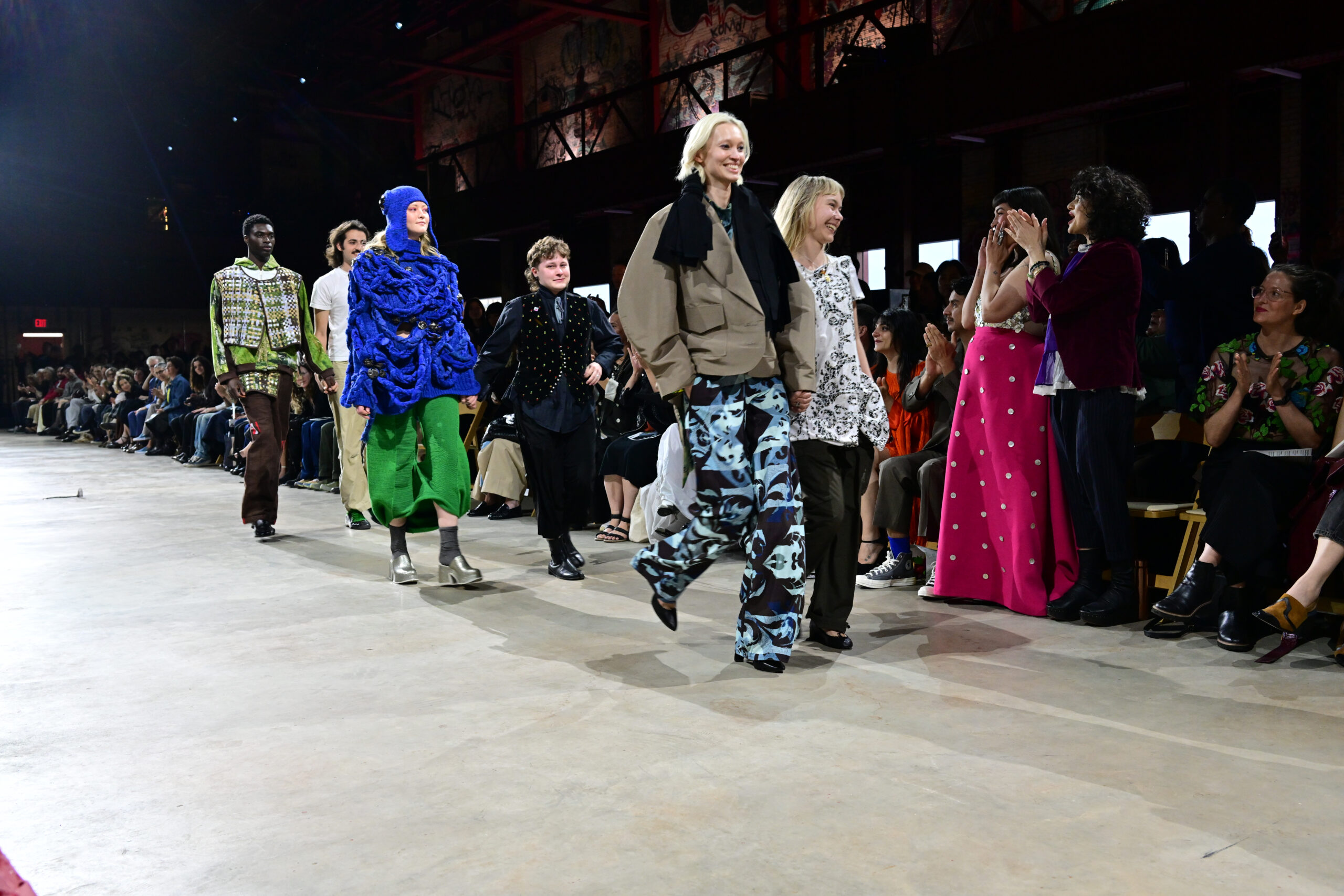 Models walk down the runway during a fashion show, showcasing a variety of colorful and unique outfits. The audience, seated on either side, looks on with interest and applause. The setting is industrial with exposed structures and large windows, and the atmosphere is lively and celebratory.