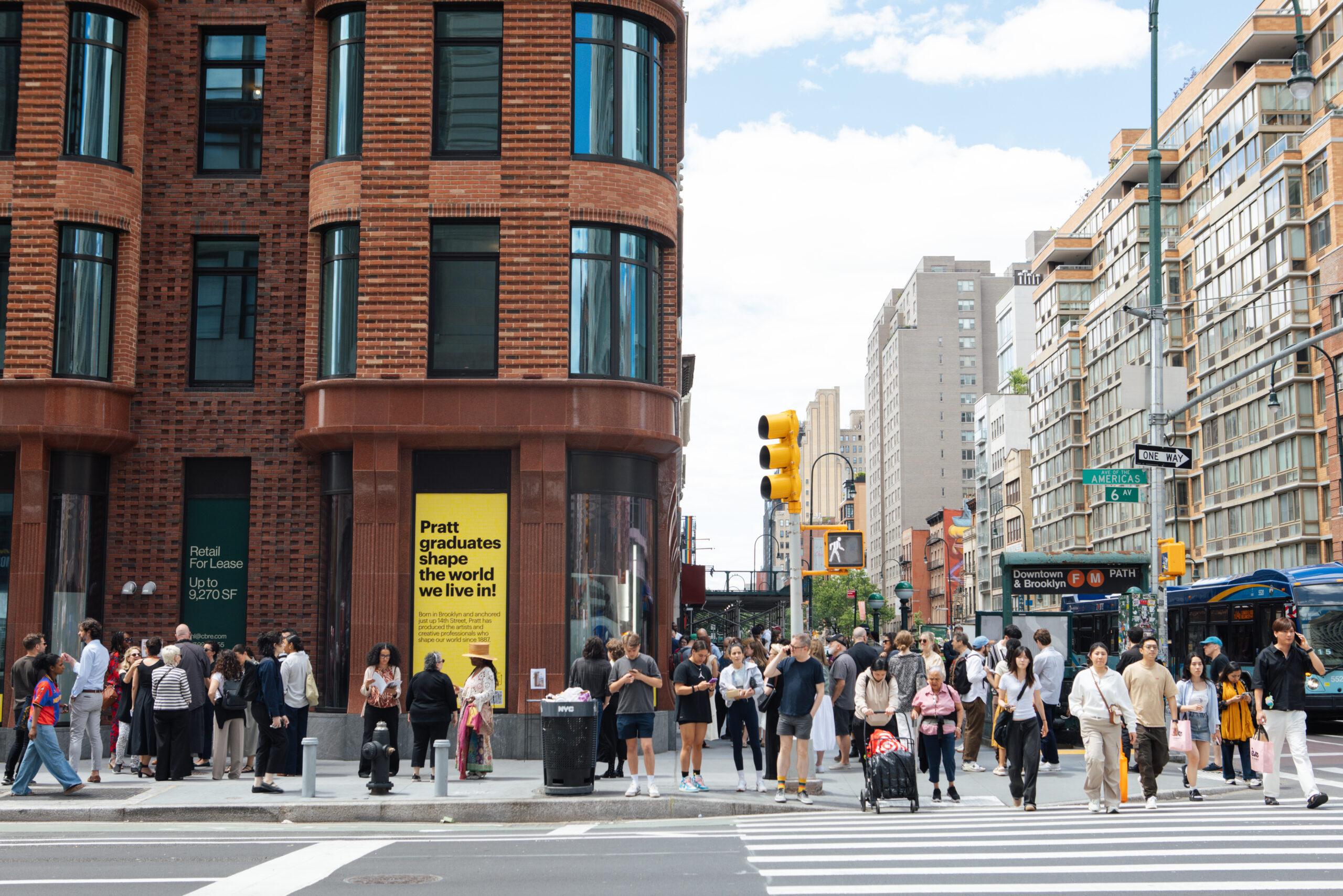 A busy urban street scene with a mix of pedestrians, including groups and individuals crossing the street. A modern brick building features a large advertisement promoting Pratt Institute. Traffic lights and street signs indicate directions for vehicles. The atmosphere is lively with a partly cloudy sky and various people engaged in conversation or using their phones.