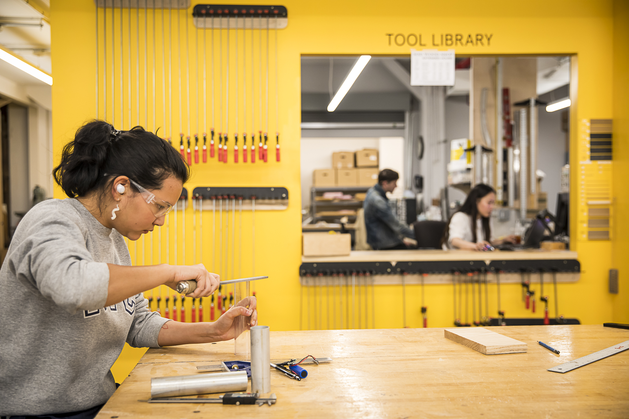 A student wearing safety goggles and earplugs at work in a workshop. Tools are around them. A Tool Library where they can check out tools is in the background.