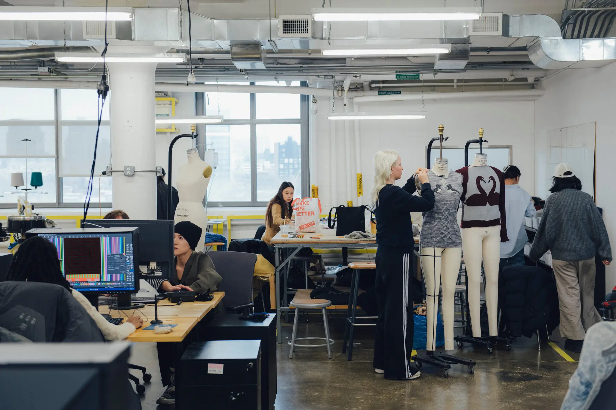 A spacious fashion design studio with students actively working at different stations. Some students are seated at computer desks analyzing color-coded digital patterns, while others are draping and styling garments on dress forms. The room is filled with mannequins, sewing equipment, and worktables, with large windows providing natural light and a view of an urban skyline in the background. The environment reflects a dynamic, hands-on learning space.