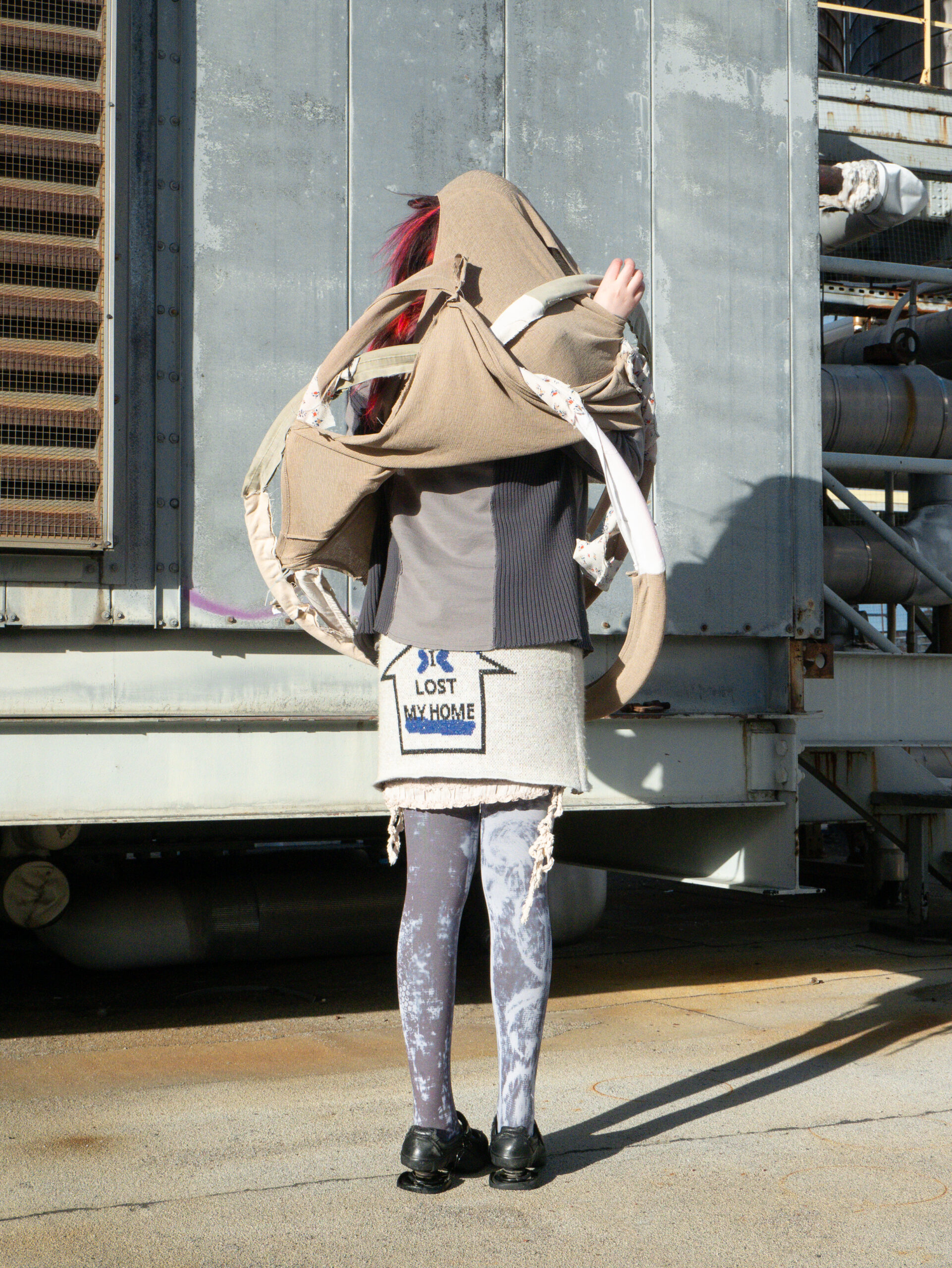 A person stands with their back to the camera in front of a large industrial metal wall. They wear an unconventional outfit consisting of layered and deconstructed garments, including a gray textured jacket, a wool skirt with a patch reading 