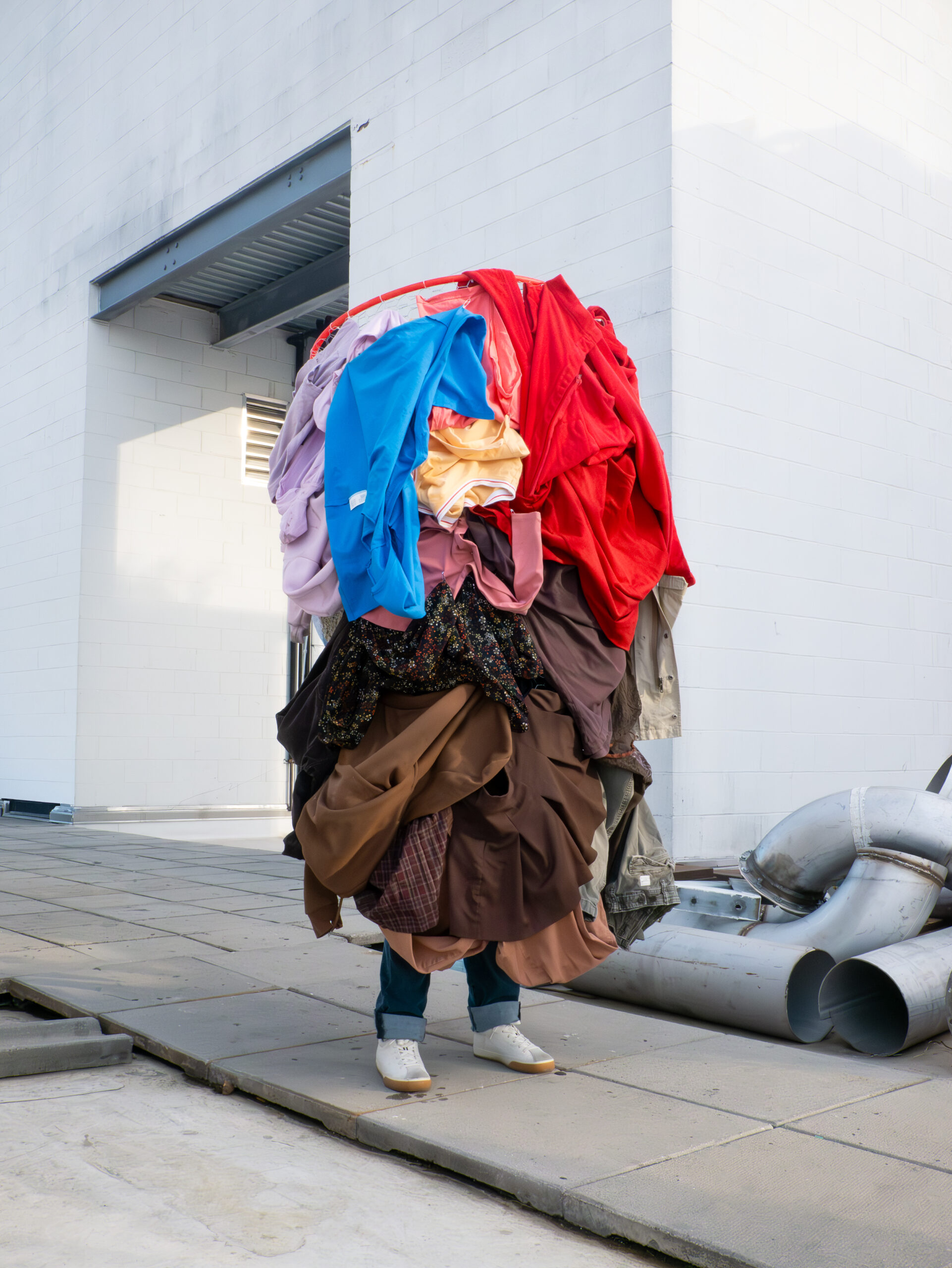 A person stands outside a modern industrial building, completely concealed by an enormous, multi-layered pile of colorful clothing balanced on their head and body. Only their shoes and lower legs are visible beneath the mound of garments, which include reds, blues, browns, and florals. The scene presents a sculptural, surreal commentary on fashion, waste, or identity.