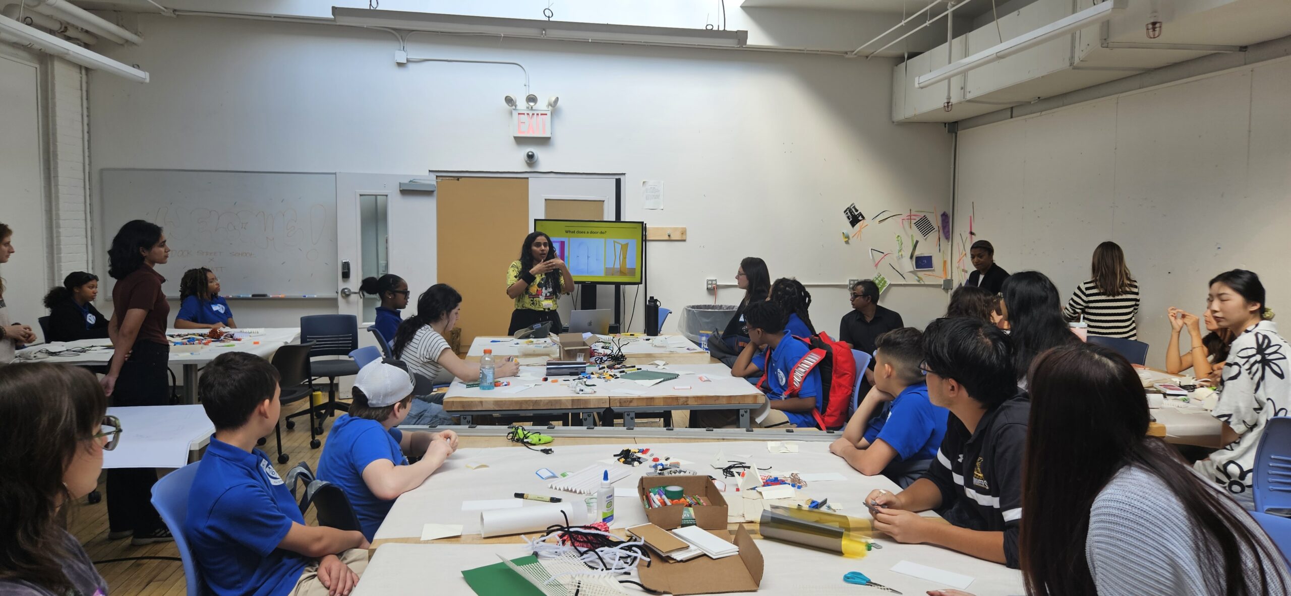 A workshop in a classroom setting, with participants seated at tables and engaged in a project. In the foreground, a diverse group of students listens attentively to a presenter who is gesturing towards a screen displaying a slide titled "What does a door do?" Various craft supplies are scattered on the tables. The room features bright lighting and walls decorated with colorful artwork.