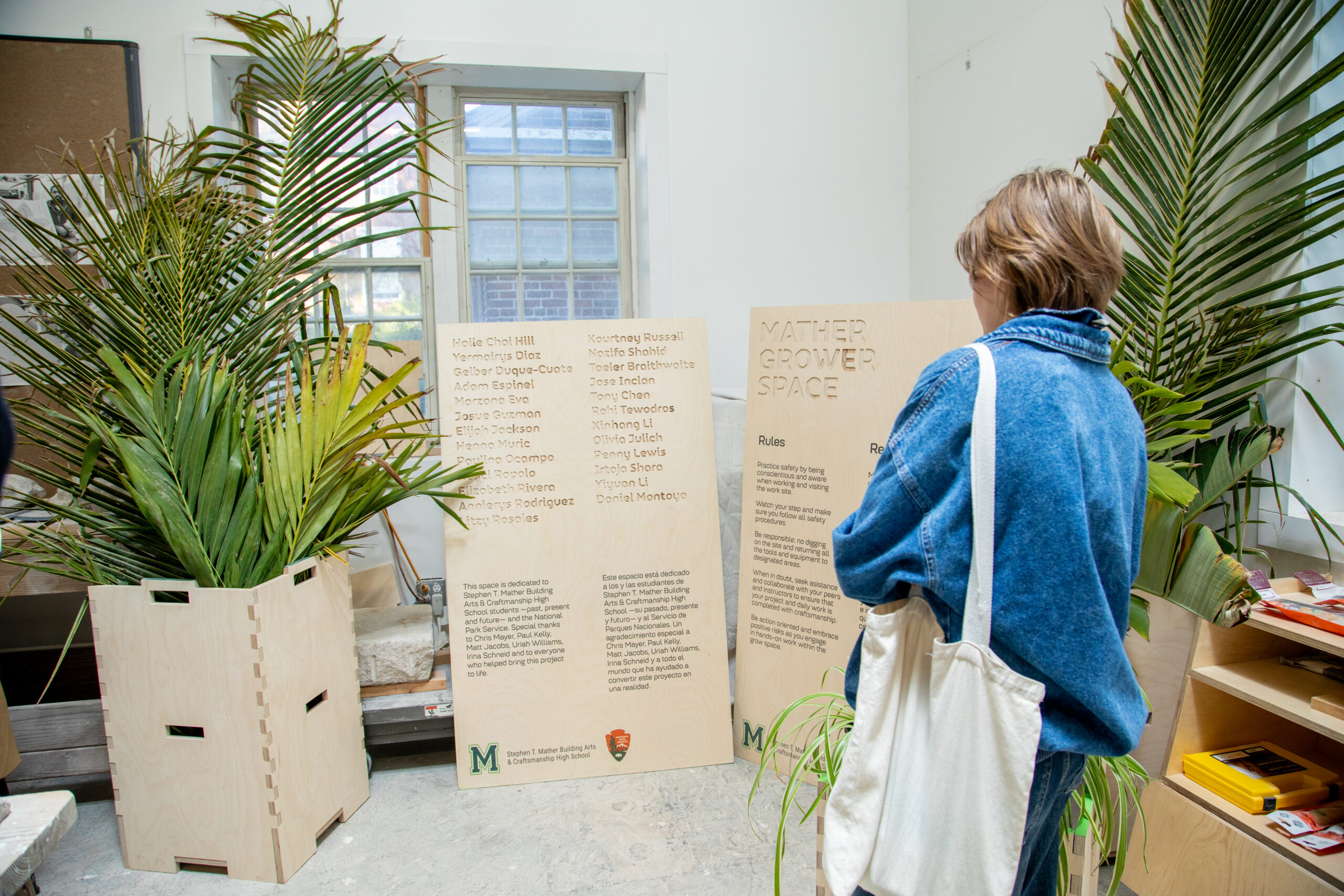 A modern workshop interior features three individuals engaging with tools and materials. One person sits on a stool at the back, focused on a laptop. Two others stand at a workbench: one man, wearing headphones, is inspecting tools, while a woman, dressed in a tank top and jeans, appears to be in conversation. The space is well-lit with large windows, showcasing a green landscape outside, and equipped with wooden cabinetry and work surfaces.