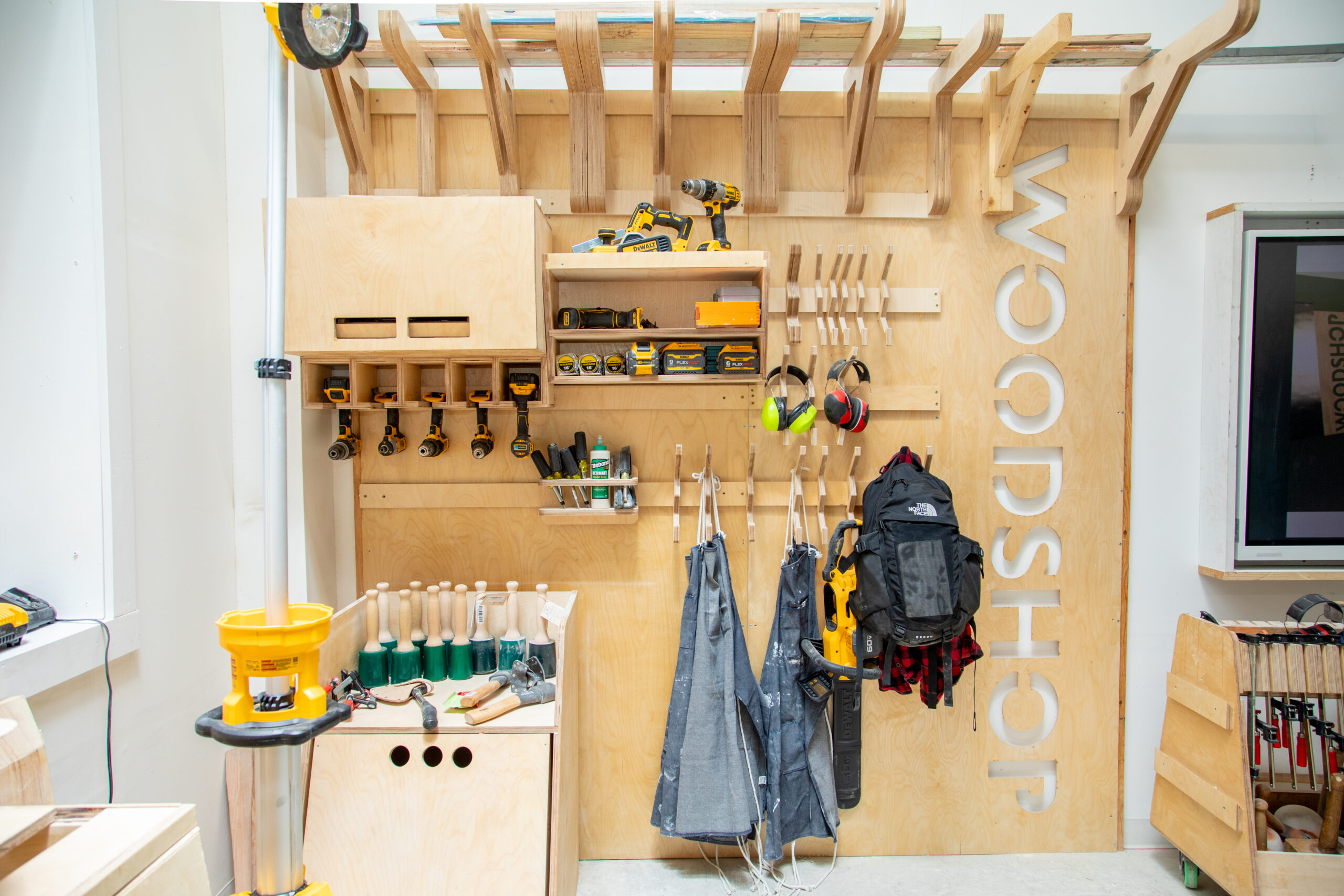 A modern workshop is depicted with light wood finishes and green accents. Three individuals are present: one person is seated at a workbench, facing away, while another stands at the center, wearing headphones and looking down, and a third person is engaged in conversation. Various tools and supplies are visible on the workbenches, and large windows provide natural light and a view of greenery outside.