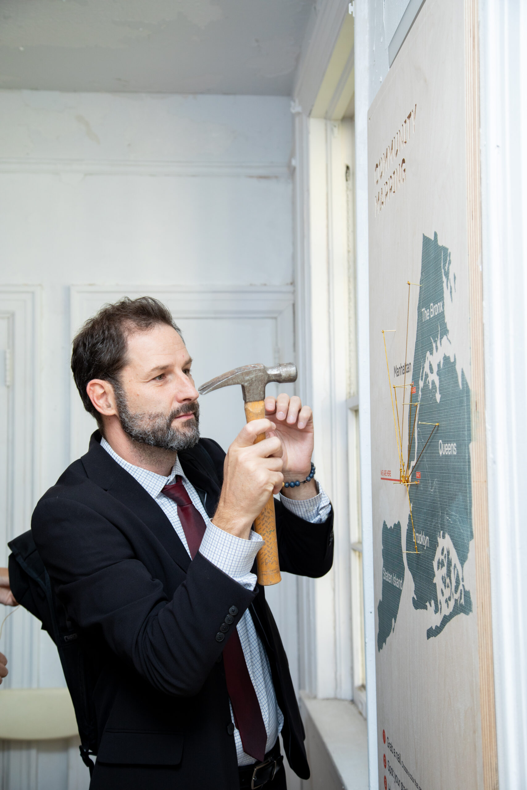 A wooden wall displays quotes and images of students involved in a collaborative design project. There are hooks with headphones and a paint-splattered apron hanging. Text from Annieyrs Rodriguez, Ella Lunghi, and Henna Muric reflects on their experiences and learnings from working together and considering various perspectives in design. The overall aesthetic combines natural wood with graphic imagery and text.