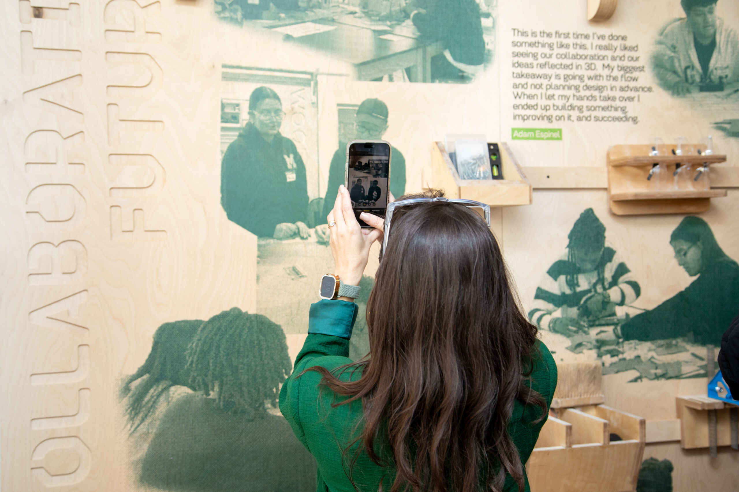 A wooden wall display features images of students engaged in design-related activities, with quotes from Annjers Rodriguez, Ella Lunghi, and Henna Muric reflecting their experiences and thoughts on collaboration and creativity. Below the images, there are wooden hooks holding a paint-splattered apron and headphones, enhancing the interactive and educational atmosphere of the space.