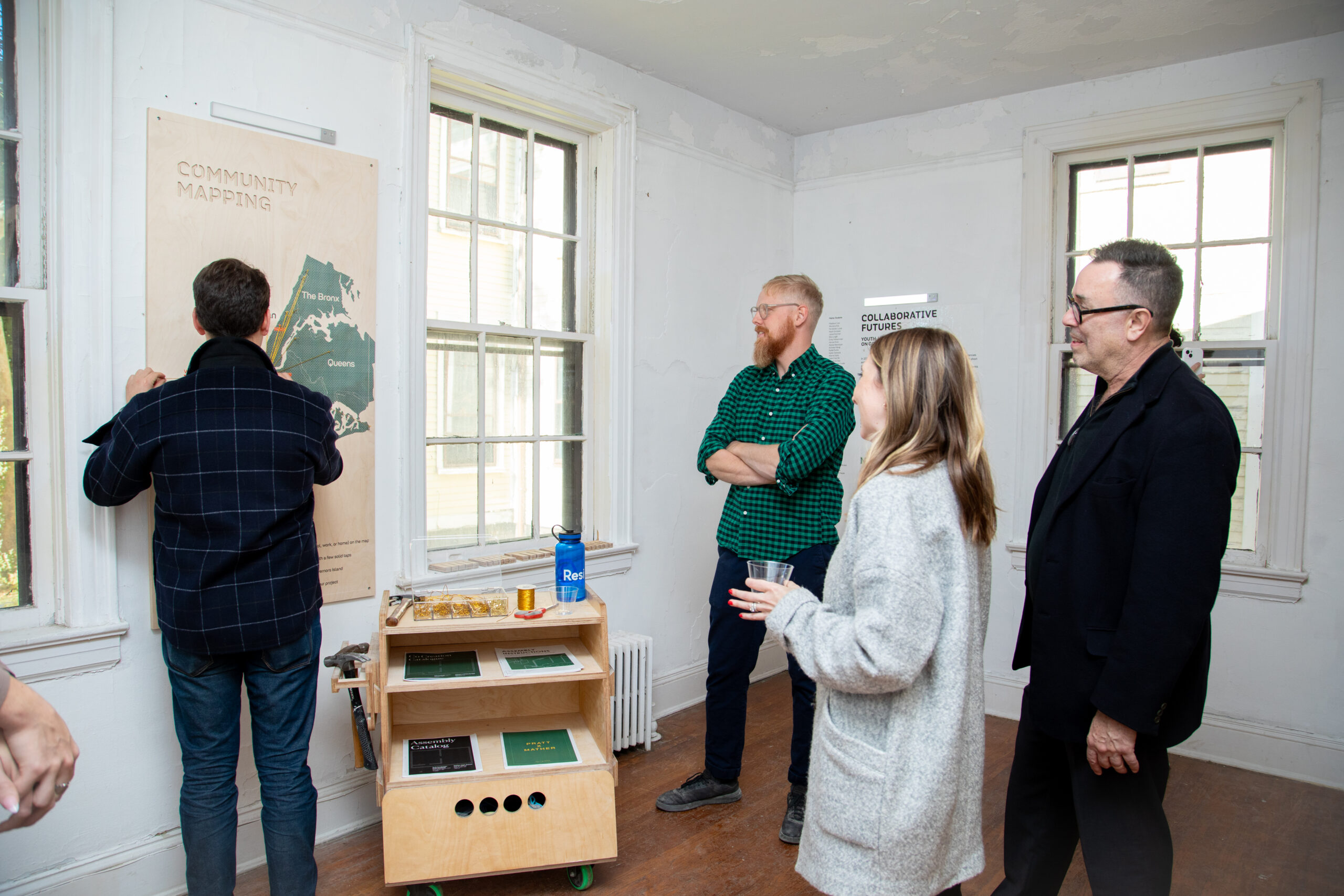 A wooden wall displays text and images from students at Mather, featuring three quotes highlighting their experiences and reflections on collaborative design work. Below the text, there are hooks with a pair of headphones and a blue apron hanging. Images of students engaged in hands-on activities are integrated into the background, creating a communal and creative atmosphere.