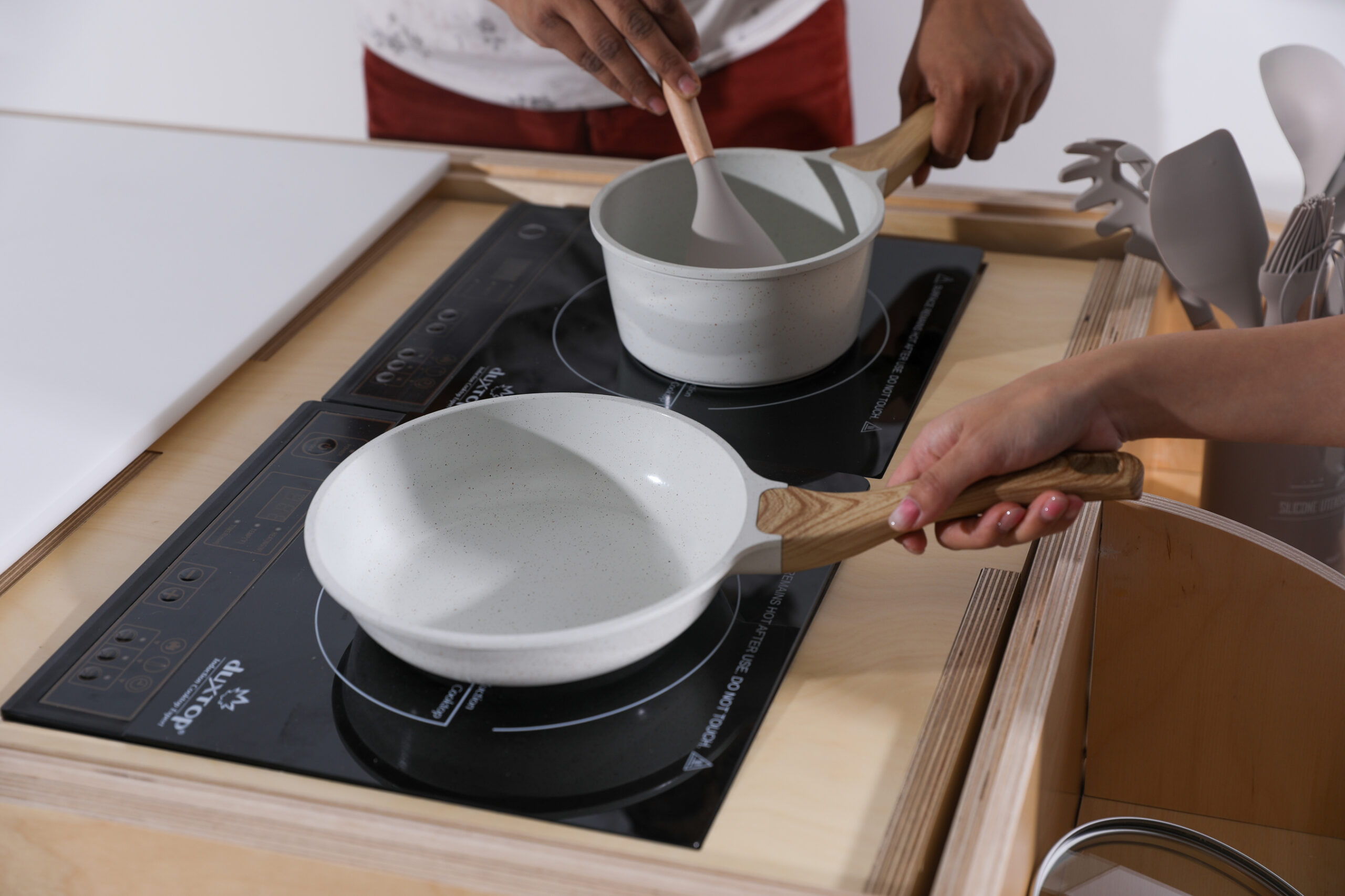 A wooden mobile cart with a rounded design featuring red accents. The cart includes various storage compartments with drawers and open sections. It has large bicycle-like wheels for mobility and is equipped with kitchen utensils displayed in a container on top. People are standing nearby, observing the cart in a classroom setting with a carpeted floor.