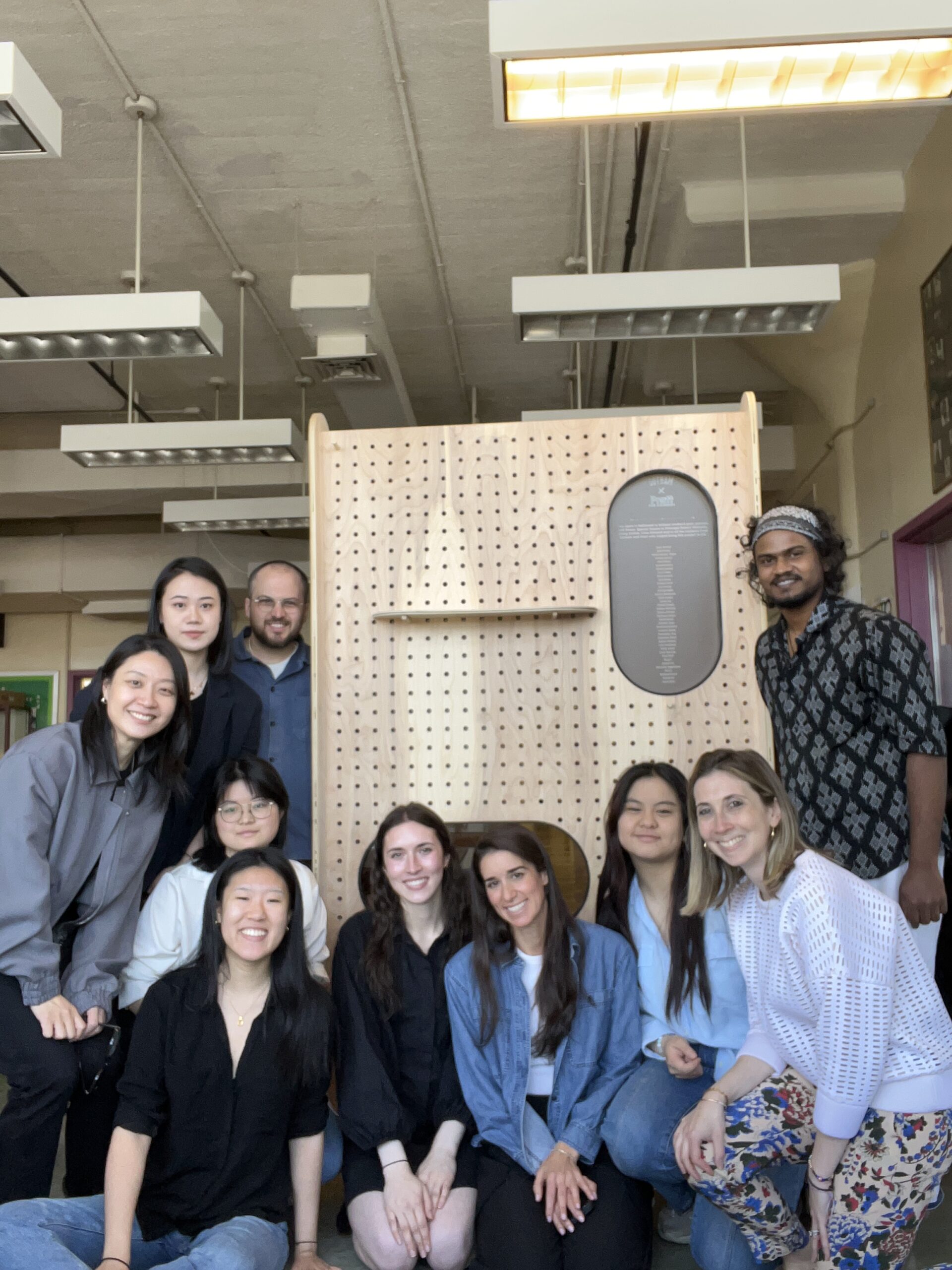 A diverse group of eleven people poses together in a well-lit indoor space. They are smiling and sitting or standing around a wooden structure with perforations. The background features overhead lights and a wall with framed pictures. The group displays a mix of clothing styles, ranging from casual to semi-formal.