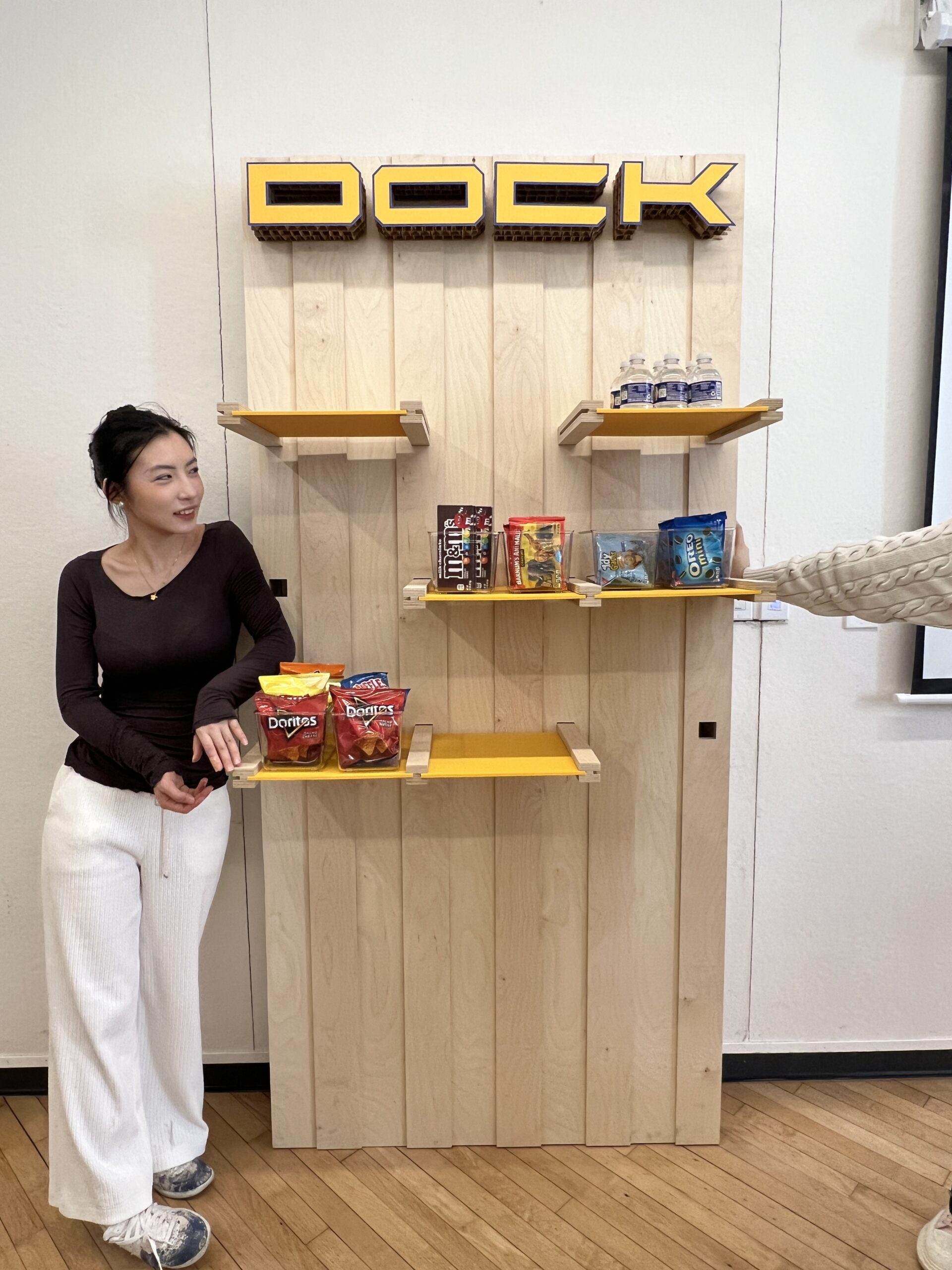 A brightly colored shop interior featuring yellow walls and shelves stocked with snacks and beverages. A teenager in a red hoodie walks past, while two girls chat nearby, one sitting on a yellow stool. The atmosphere is vibrant and playful, with a poster on the wall labeled "THE BOOK" displaying various names.