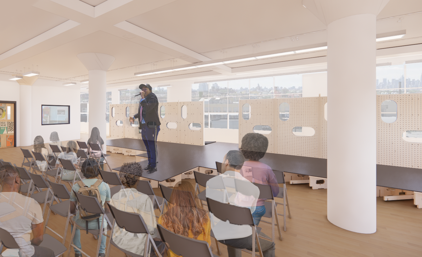 A speaker stands on a small stage in a well-lit room, addressing an audience seated in chairs arranged in rows. The audience consists of several individuals with varying hairstyles and outfits, all facing the speaker. In the background, there are wooden partitions with cut-out windows, and large windows offer a view of the city skyline outside.