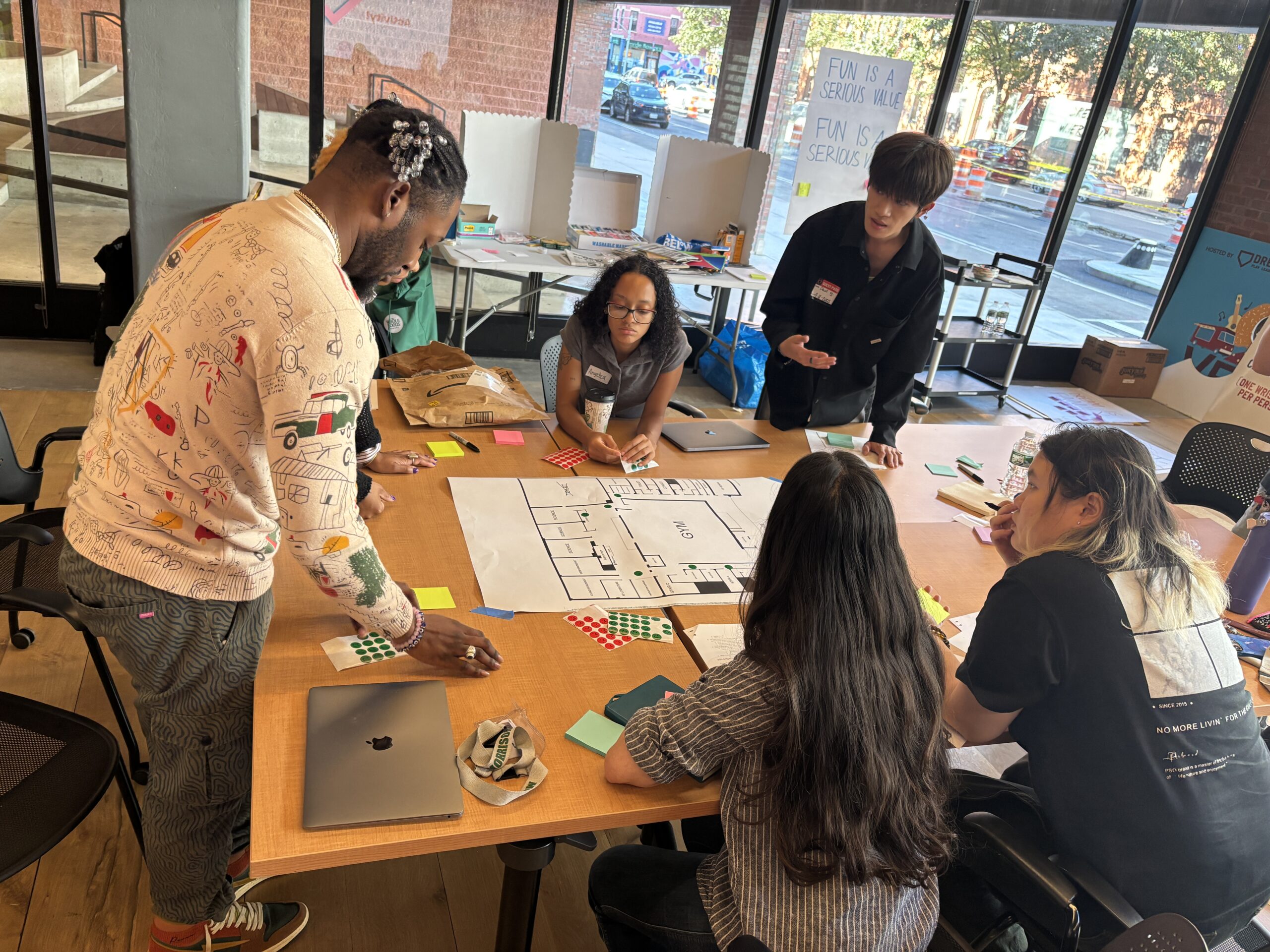 A group of six people is engaged in a collaborative activity at a wooden table covered with notebooks, sticky notes, and a laptop. One person is standing and explaining something while others are sitting, observing, or working on the table. There are colored stickers and a map-like layout drawn on paper in the center. Large windows in the background show an outdoor scene.