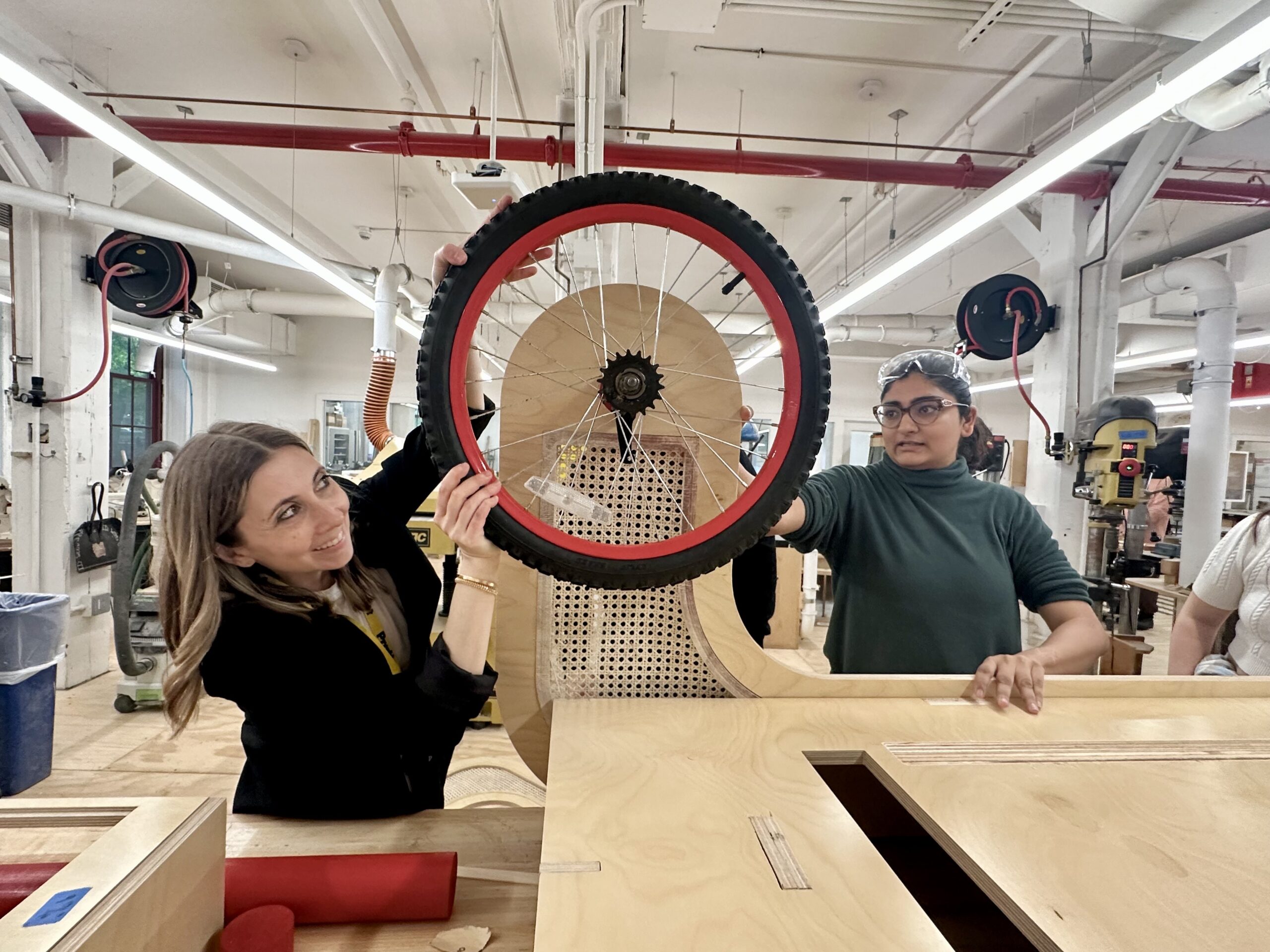 Two women are in a workshop holding up a bicycle wheel. One woman, smiling, is on the left, while the other, wearing glasses and a green sweater, holds the wheel on the right. The background features woodworking equipment, including a large wooden structure and tools, under bright overhead lights.