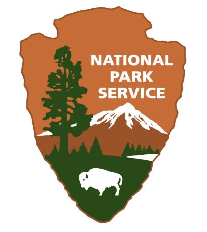 The National Park Service emblem in the shape of an arrowhead. It features a large tree, mountains, water, and a bison with the words 