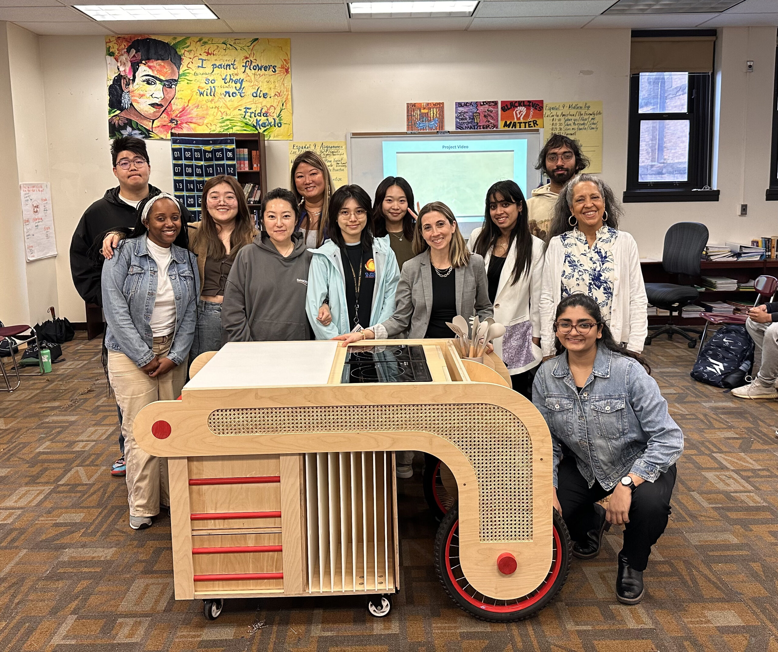 A group of diverse individuals stands together in a classroom, posing around a custom-designed wheeled cart with kitchen equipment. The cart features wooden shelves, red accents, and kitchen utensils visibly placed on top. Behind them, colorful artwork and posters decorate the walls. A projector screen displays a presentation, and additional students are seen in the background.