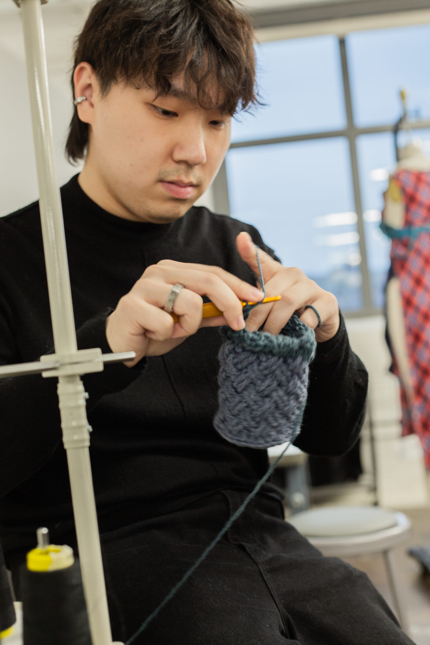 A person with short dark hair is focused on crocheting in a bright, modern workspace. They are wearing a black turtleneck and holding a crochet hook, working on a textured piece made of yarn. Various sewing materials and garments are visible in the background, indicating a creative environment. Natural light streams in through a large window.