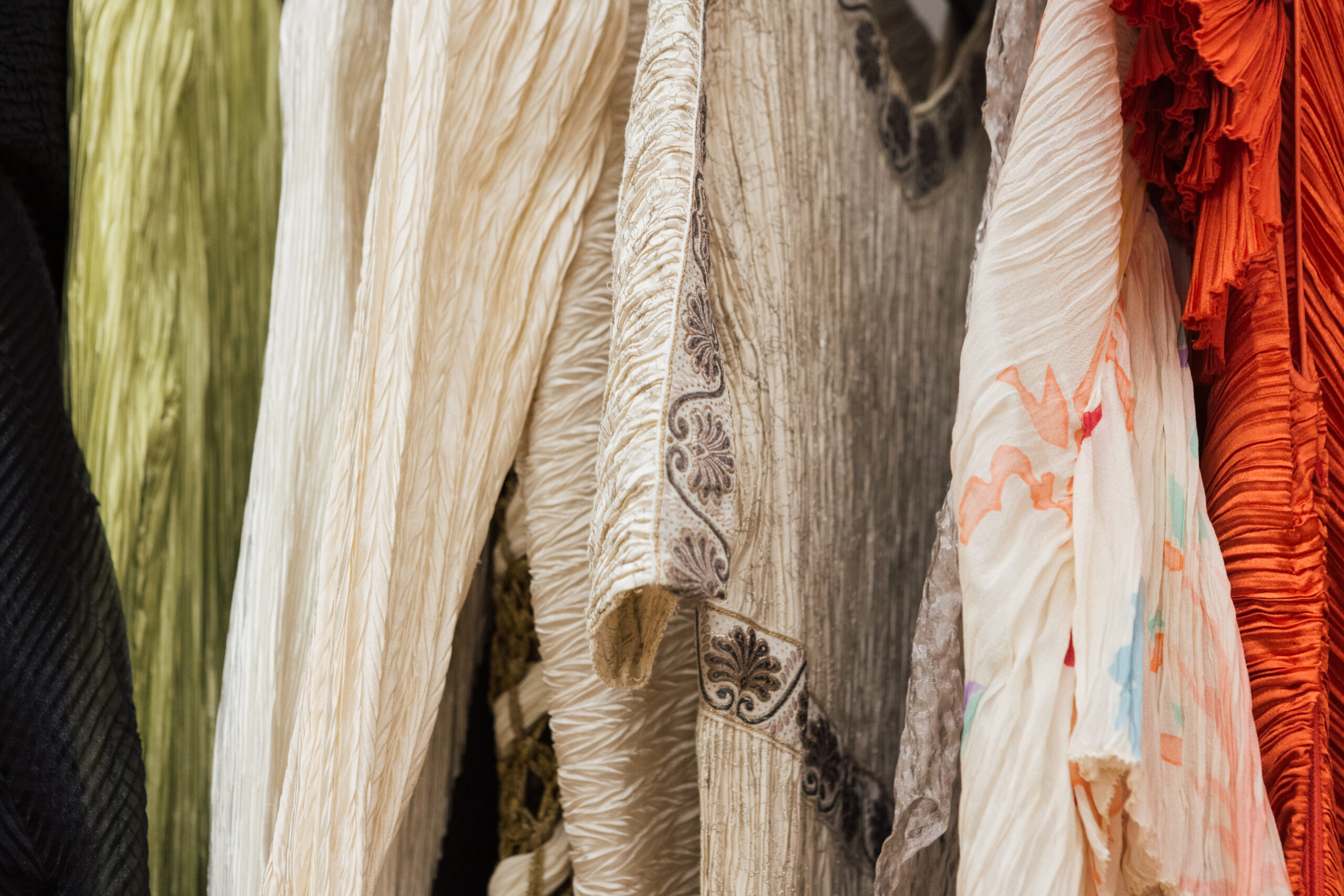 Close-up of a collection of textured garments hanging together. The fabrics feature various colors and patterns, including green, ivory, light gray, white with floral designs, and vibrant orange. Some fabrics have pleats or intricate embellishments, showcasing different styles and materials.