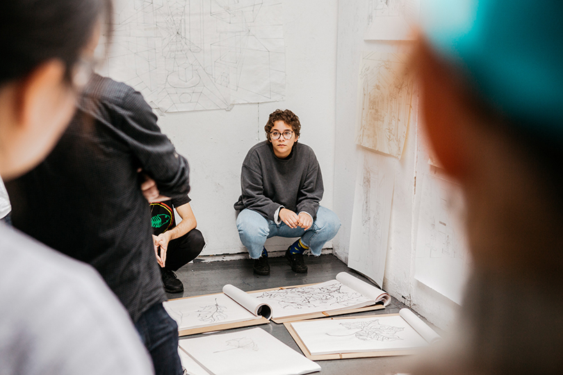 A photograph of a student crouching near open sketchbooks laid out on the floor. There are pieces of sketch paper pinned up on the wall around them. A group of students is looking towards the crouching student.