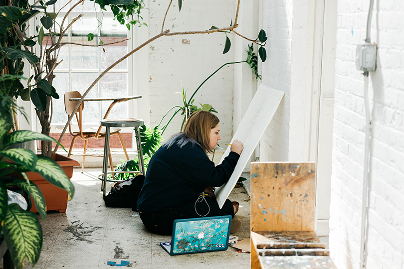 Student working on art in a bright room, the brick walls are painted white and there is a window letting in lots of sunlight. There are plants all around.