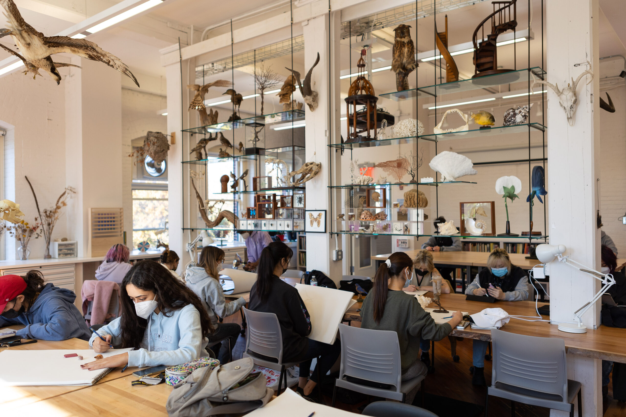 Photograph of the Foundations Lab showing large work tables and study models on shelves. Students sit at the tables working with large sketchbooks.