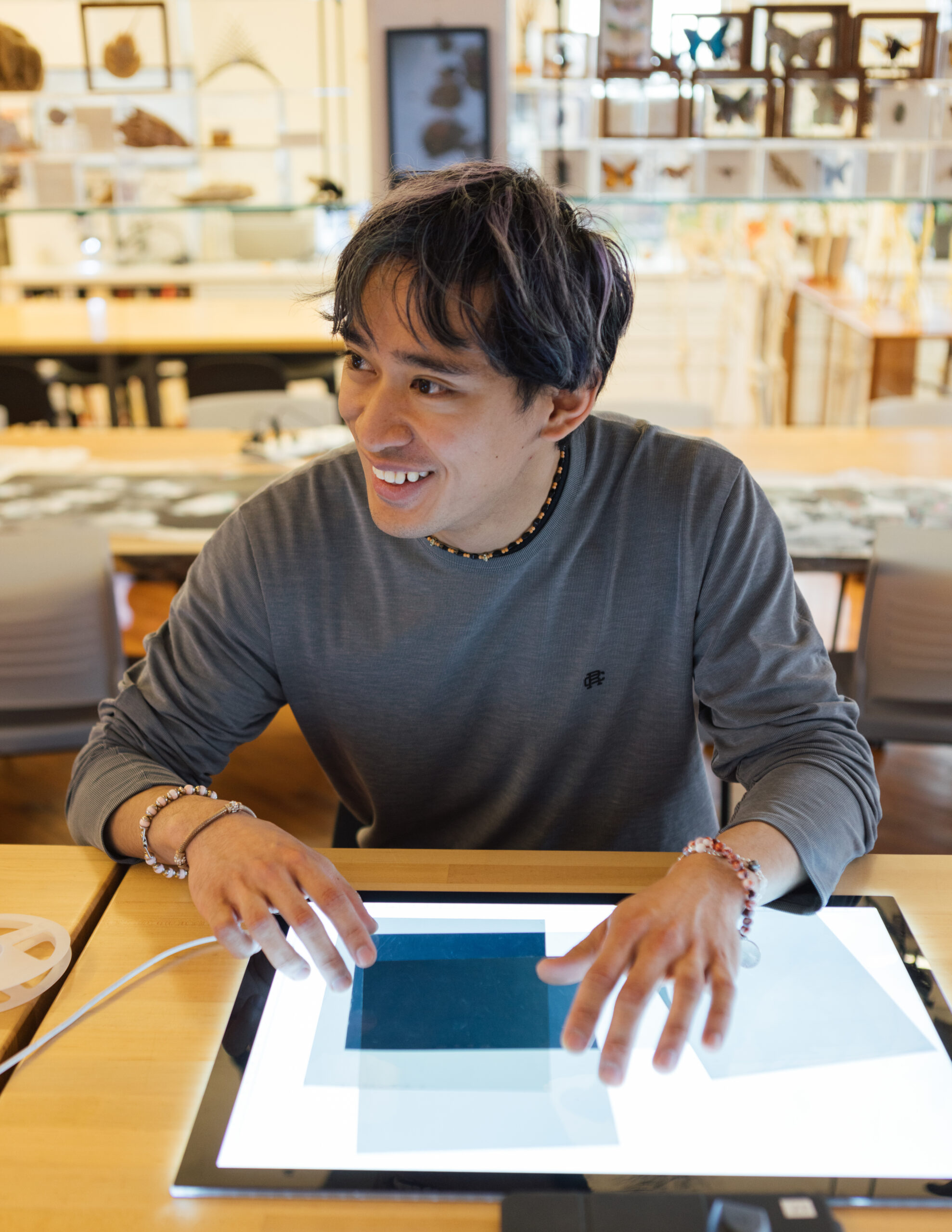 student, working on tablet, in large lab room, many artworks on wall in background