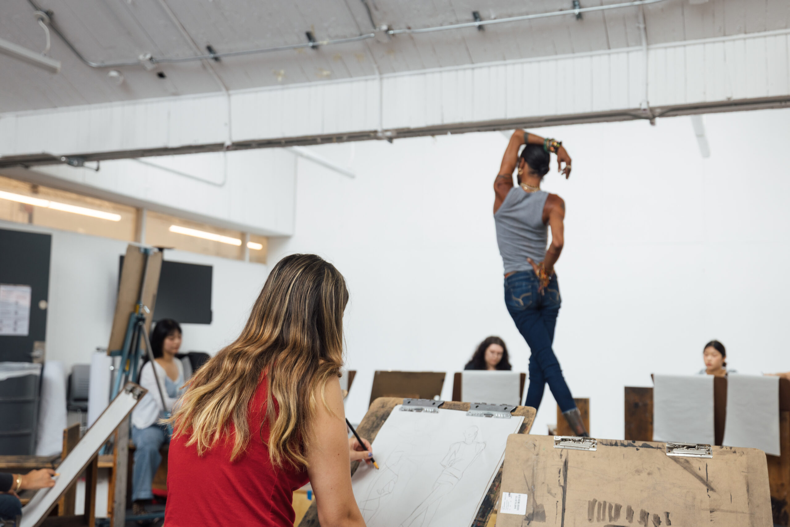 Students participate in a figure drawing session inside a large studio classroom. One student sketches on a drawing board in the foreground while a standing model poses on a platform at the front of the room. Additional students are seated at easels in the background, observing and drawing in a well-lit academic art studio.