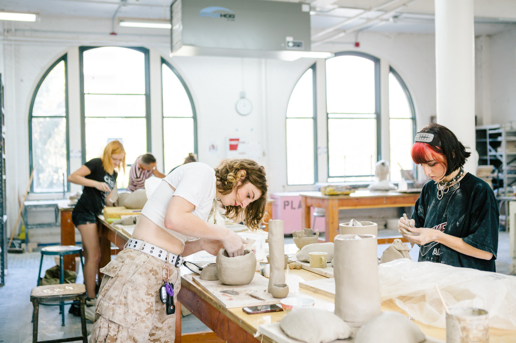 A bright pottery studio with large arched windows that let in ample natural light. Several students are focused on their ceramic projects, shaping and molding clay on work tables filled with clay pieces in various stages of completion. The foreground shows two students intently working on their clay pieces, one with curly hair and a white top, and the other with a red and black hairstyle, wearing a black oversized shirt. The studio atmosphere is creative and collaborative, with tools and clay objects scattered across the tables.
