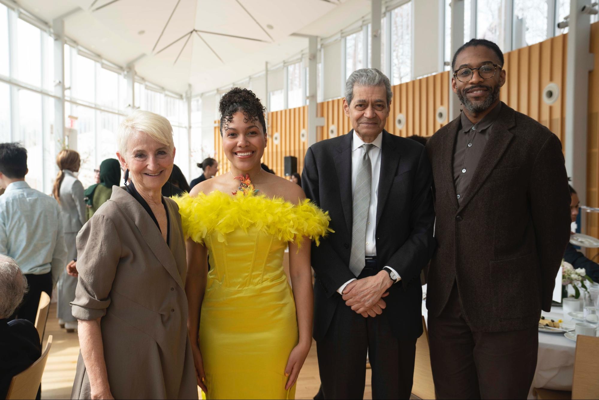 A group of four people, including a woman in a bright yellow dress, stand together smiling for a photo in a well-lit room with large windows.