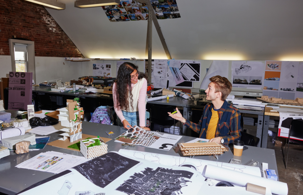 Two students working in an architecture studio, surrounded by various models, drawings, and design materials. They are engaged in a discussion, with one student pointing to a model on the table and the other holding a paper. The studio is filled with creative projects and has a bright, collaborative atmosphere.