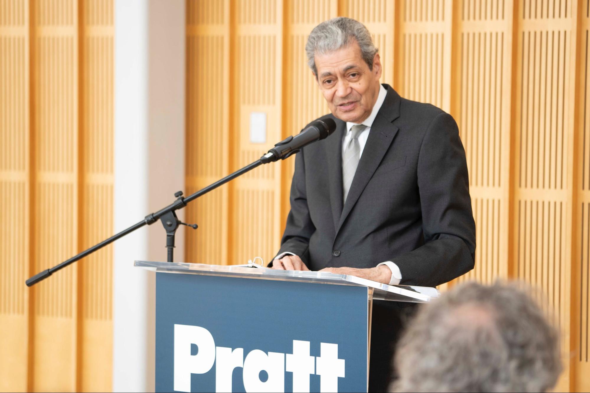 A man in a suit speaks at a podium with a "Pratt" sign, addressing an audience in a wood-paneled room.