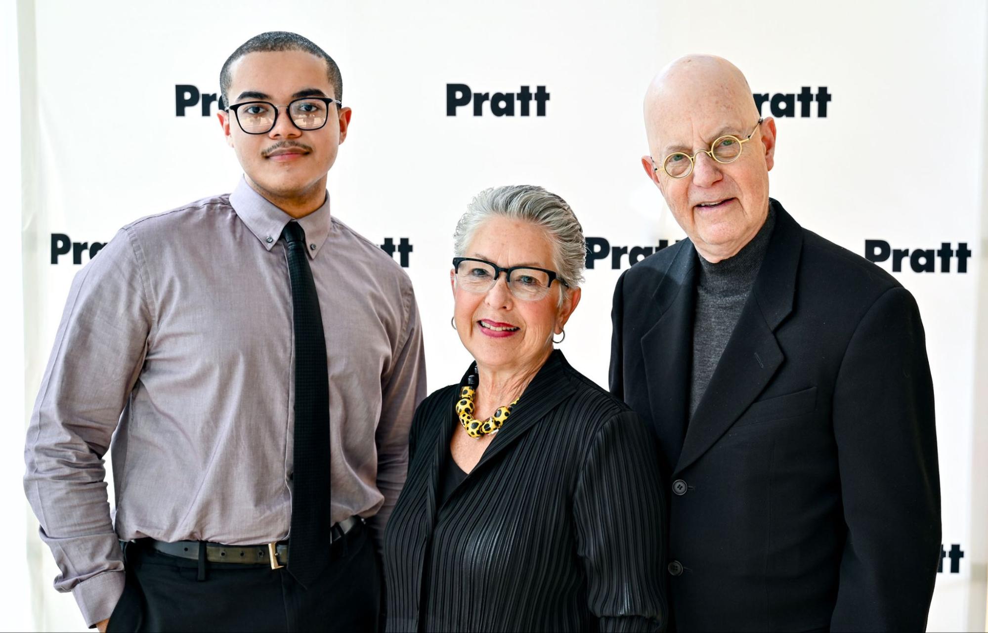 three people, standing against a pratt backdrop, with the word "Pratt" in a monogram-like pattern in the background, man wearing dress shirt and tie, smiling, woman with glasses, suit and short hair cut, smiling, older gentleman with suit, smiling in foreground