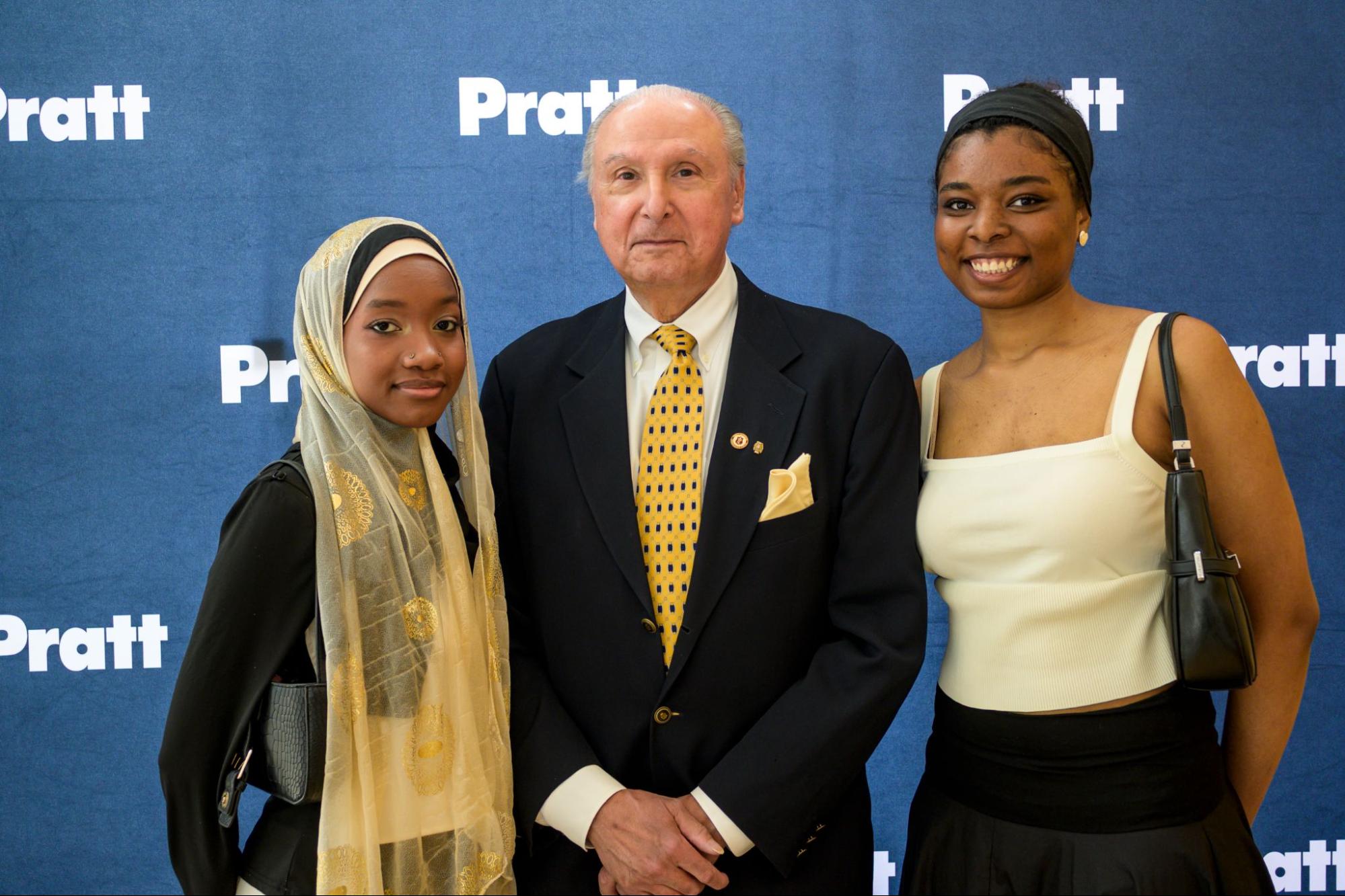 Three people pose for a formal photograph in front of a blue step-and-repeat backdrop with the white Pratt Institute logo repeated across it. In the center, an older man in a dark suit, white shirt, and yellow-patterned tie stands with his hands clasped. To his left, a young woman wears a light gold and silver patterned hijab and a black top, and to his right, another young woman in a cream-colored top with black accessories smiles at the camera. The setting appears to be a celebratory or academic event.