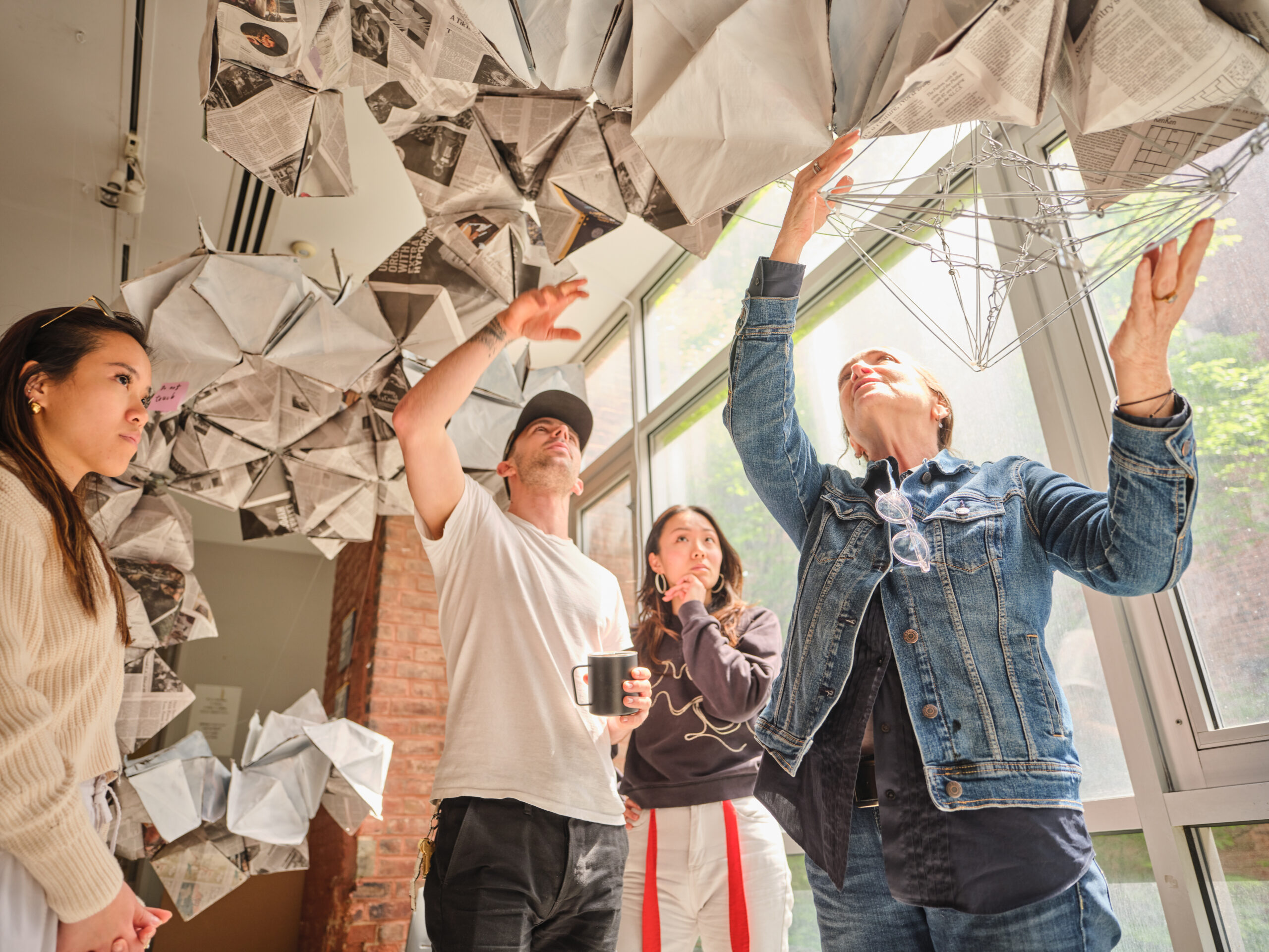 Four people stand under a hanging installation of folded paper. They are pointing and looking at different parts of the piece.