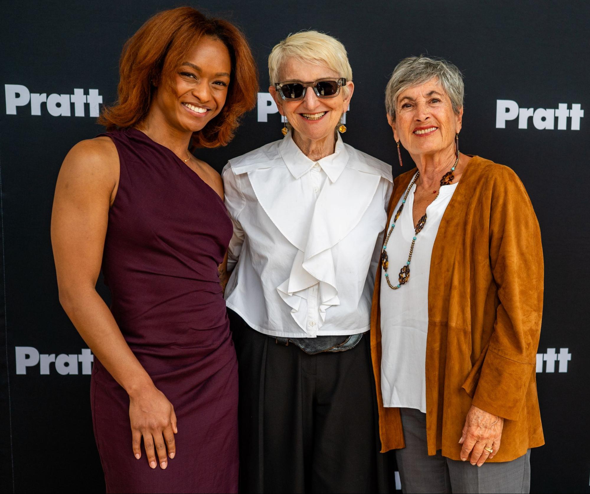 Three women stand side by side in front of a black backdrop printed repeatedly with the word “Pratt.” All three smile at the camera. The woman on the left wears a sleeveless burgundy dress. The woman in the center wears a white blouse with a ruffled front, dark pants, and sunglasses. The woman on the right wears a white top with a long necklace and a rust-colored jacket. The image appears to be taken at a formal event or reception.
