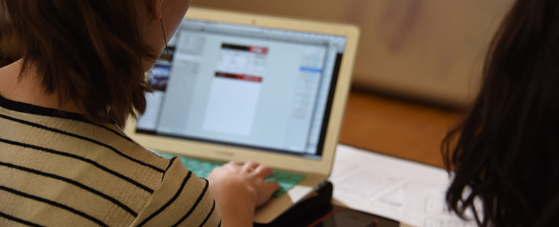 A photo of a person typing on computer taken from behind. Another person's head can be seen at the right edge of the photo