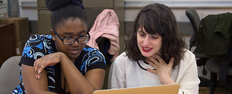 A photo of two students looking at a laptop screen taken from in front of them. They are sitting and you can only see the top half of their bodies