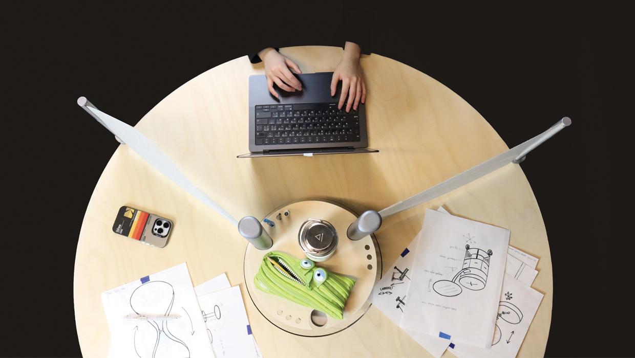 A blonde wood table with a rotating partition that creates three spaces. One space has a laptop and others hold sheets of paper.