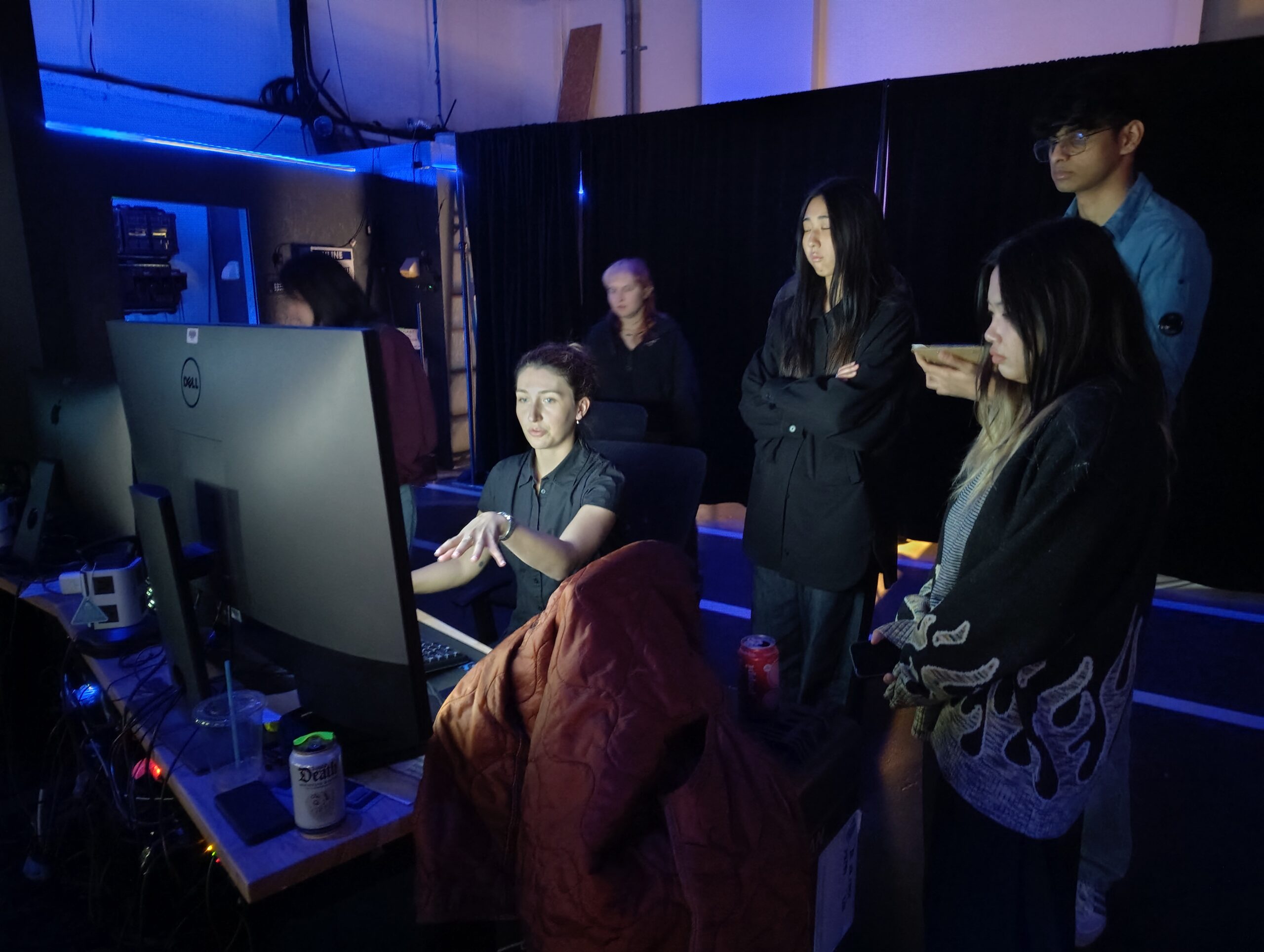 A group of five people is gathered around a large computer monitor in a dimly lit space, with blue lighting in the background. One person is seated at the computer, gesturing as they speak, while the others, standing, are observing intently. The atmosphere appears focused and collaborative, with equipment and personal items visible on the desk.