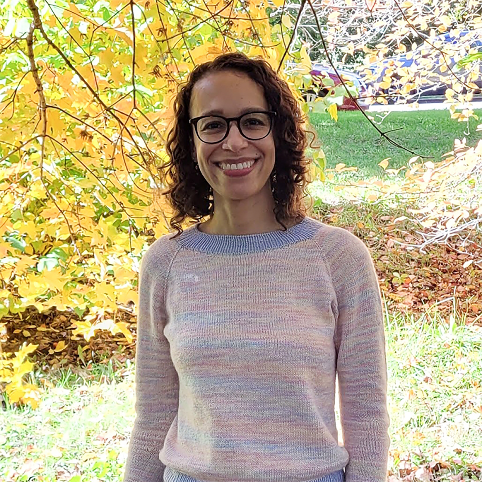 A person stands smiling in front of vibrant autumn leaves, which are various shades of yellow and orange. They have curly hair and are wearing glasses, along with a light-colored sweater. The background includes a grassy area and scattered fallen leaves.