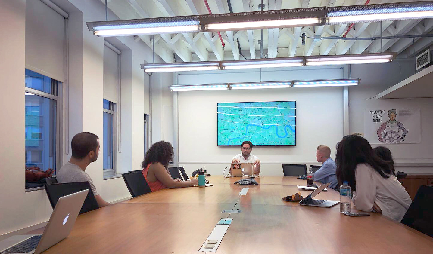 A group of students sit around a conference table watching a presenter at the front of the room. There is a screen behind the presenter with a map on it.