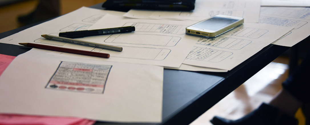 Pencils, pens, papers with hand drawn mockups, and an iPhone are on a desk.
