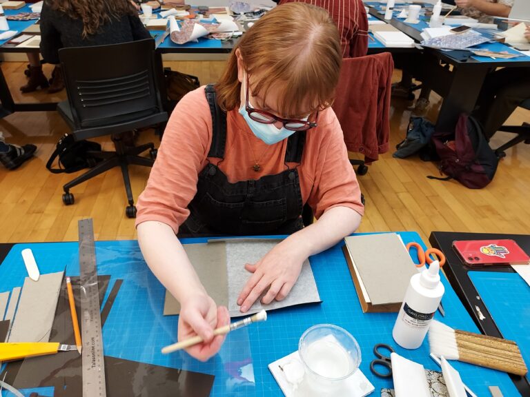 A student at a worktable with an archival document. They are holding a brush and are surrounded by their tools.