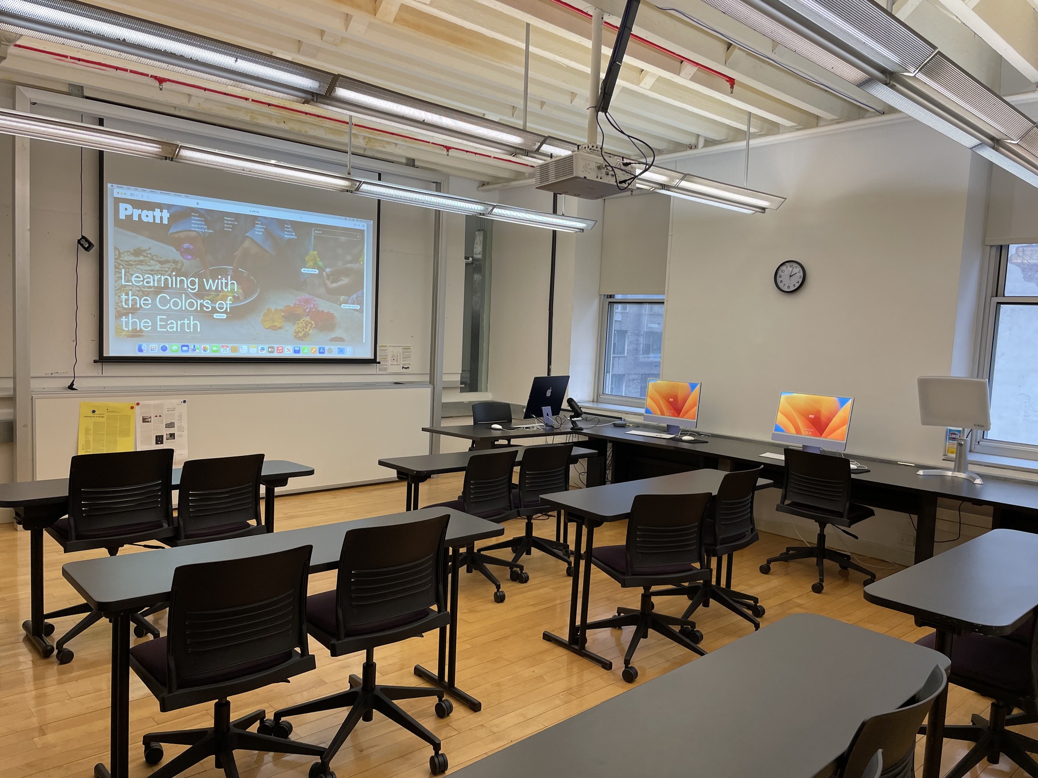 An array of work tables, some with computer work stations, others just with chairs. There is a projector screen on the wall.