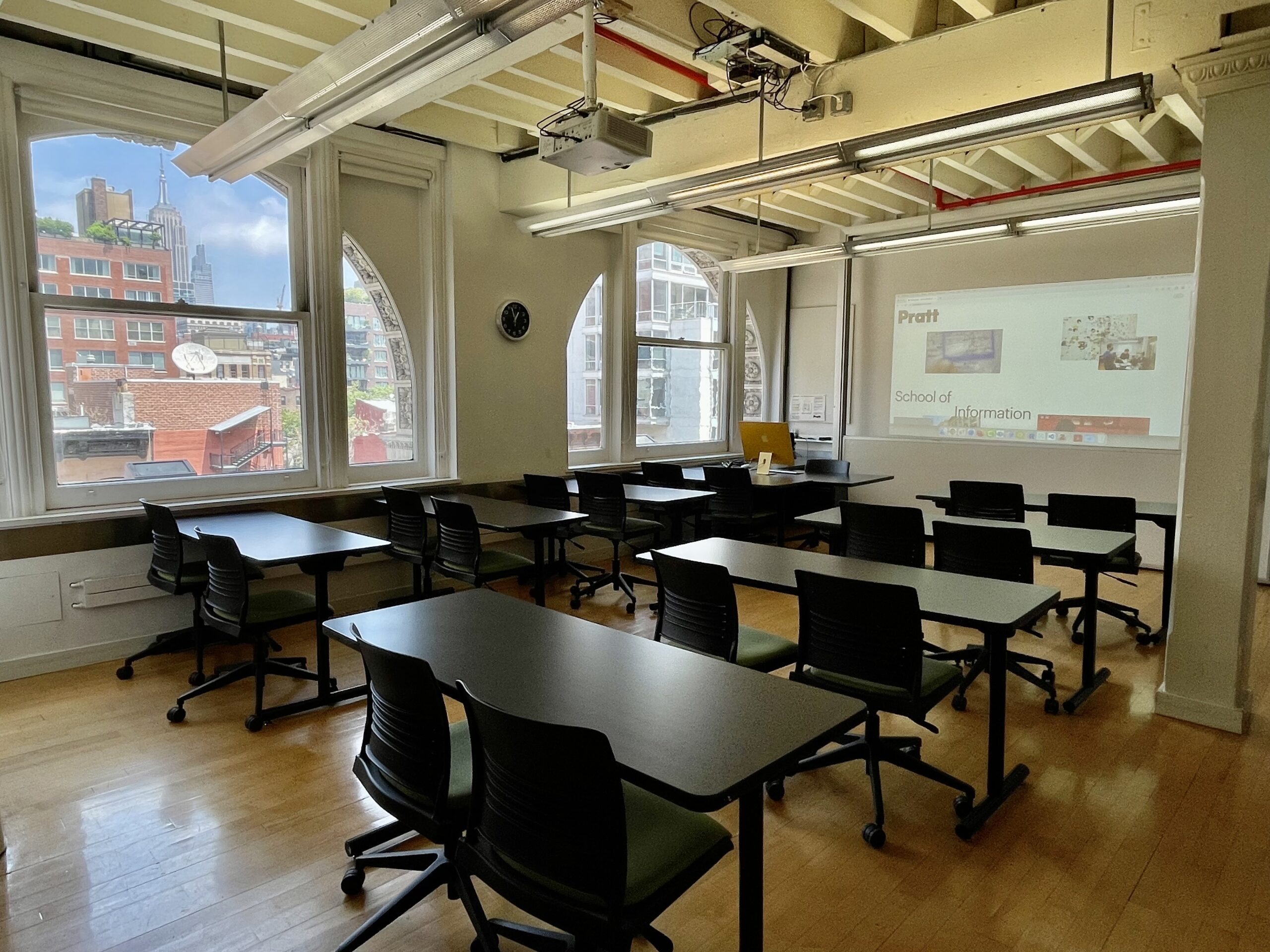8 work tables each with two chairs in a room with large windows and a projector screen.