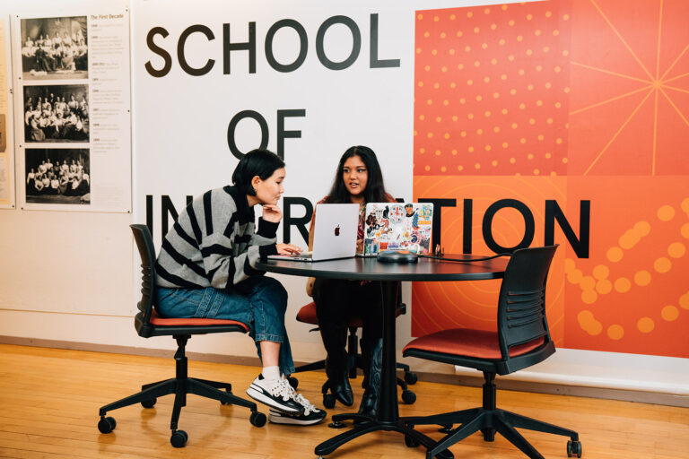 Two students sit at a communal table with their laptops. They are conversing with each other.