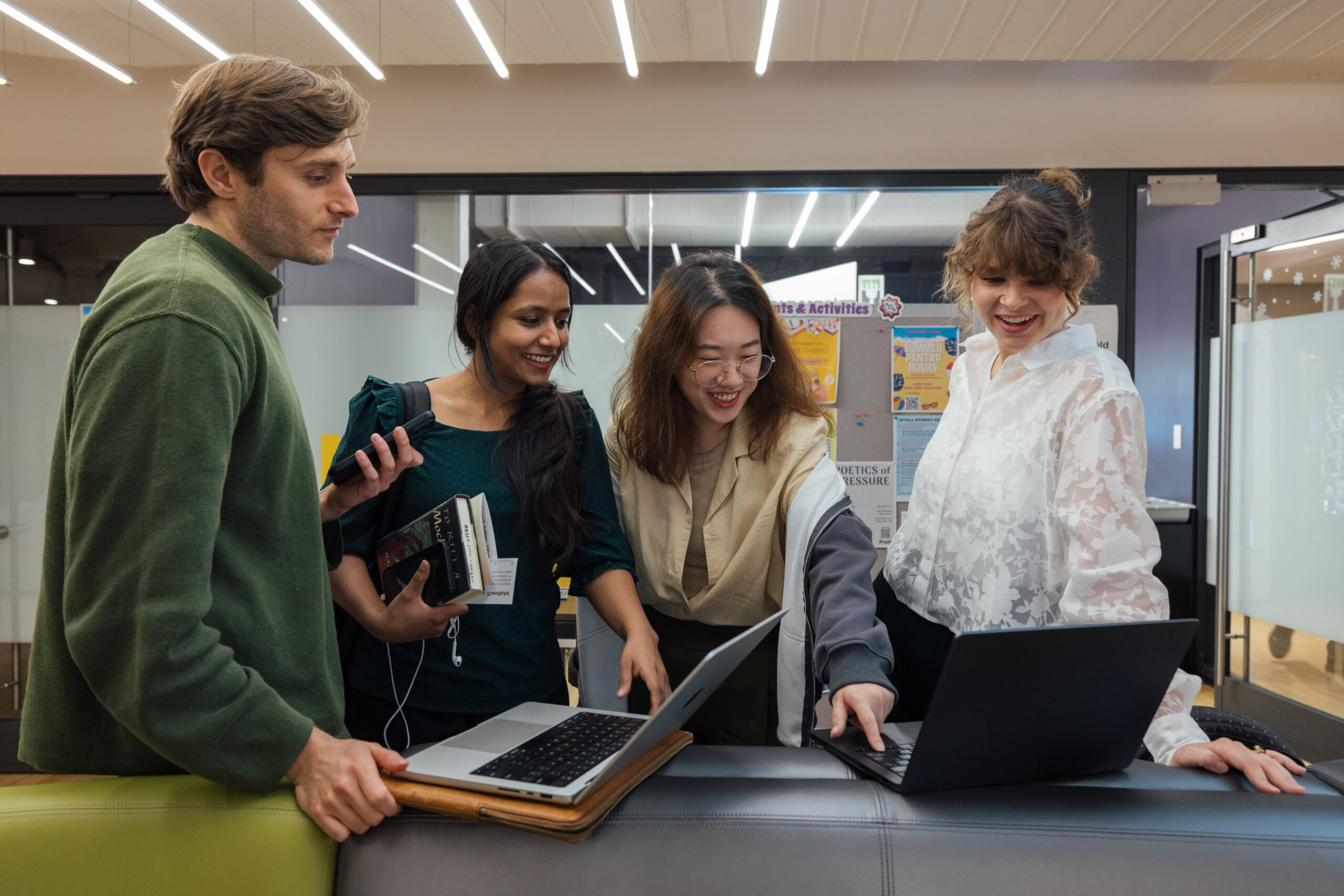 A group of four students stands together in a brightly lit common area, smiling and gathered around two open laptops. One student points at the screen while the others look on with interest. They are holding books, notebooks, and phones, indicating a collaborative academic or creative discussion. Posters and informational flyers are visible on the wall behind them.