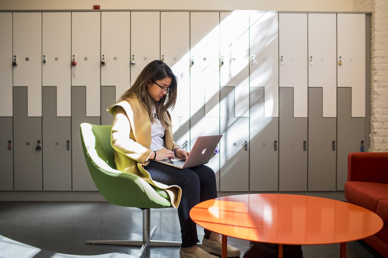 Student on campus, seated in plastic chair, in front of row of lockers, sun shining into room, causing shadows to form