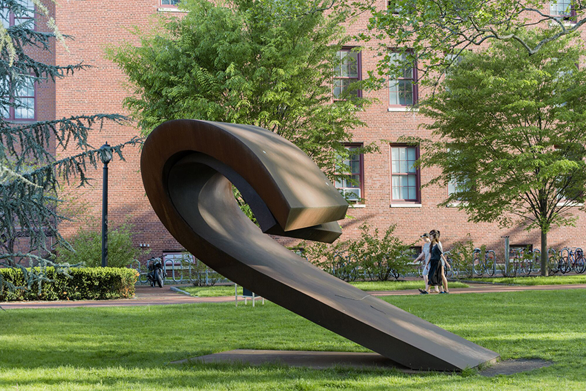 A metallic, curved sculpture rises from the ground at 40 degree angle. It is located in a lawn with a red brick building and walking students in the background