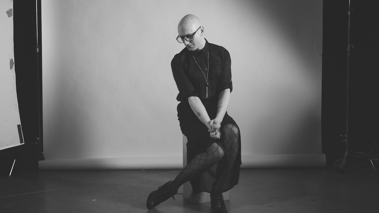 slender person with shaved head and glasses, seated on a small stool, legs bent